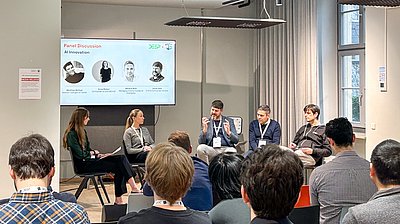 Two women and three men sit on a panel facing the audience, behind them a screen showing the topic of the discussion: AI innovation (image: Nino Strauch / Tübingen AI Center)
