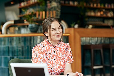 A young woman sitting at a table in an easy chair, talking to two people who are shown blurred from the back. (image: Tübingen AI Center / Nino Strauch)