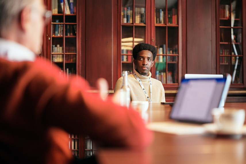 A young man in a jumper sits at a table, behind him a bookcase, in front of him a laptop and another man at the table, who is visible from behind and who is blurred (image: Tübingen AI Center / Nino Strauch)